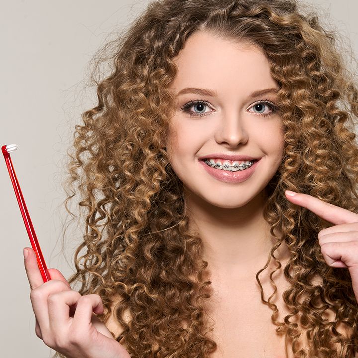 fată adolescentă, cu păr șaten și creț, ținând o periuță de dinți în mțnă și poartă aparat dentar.